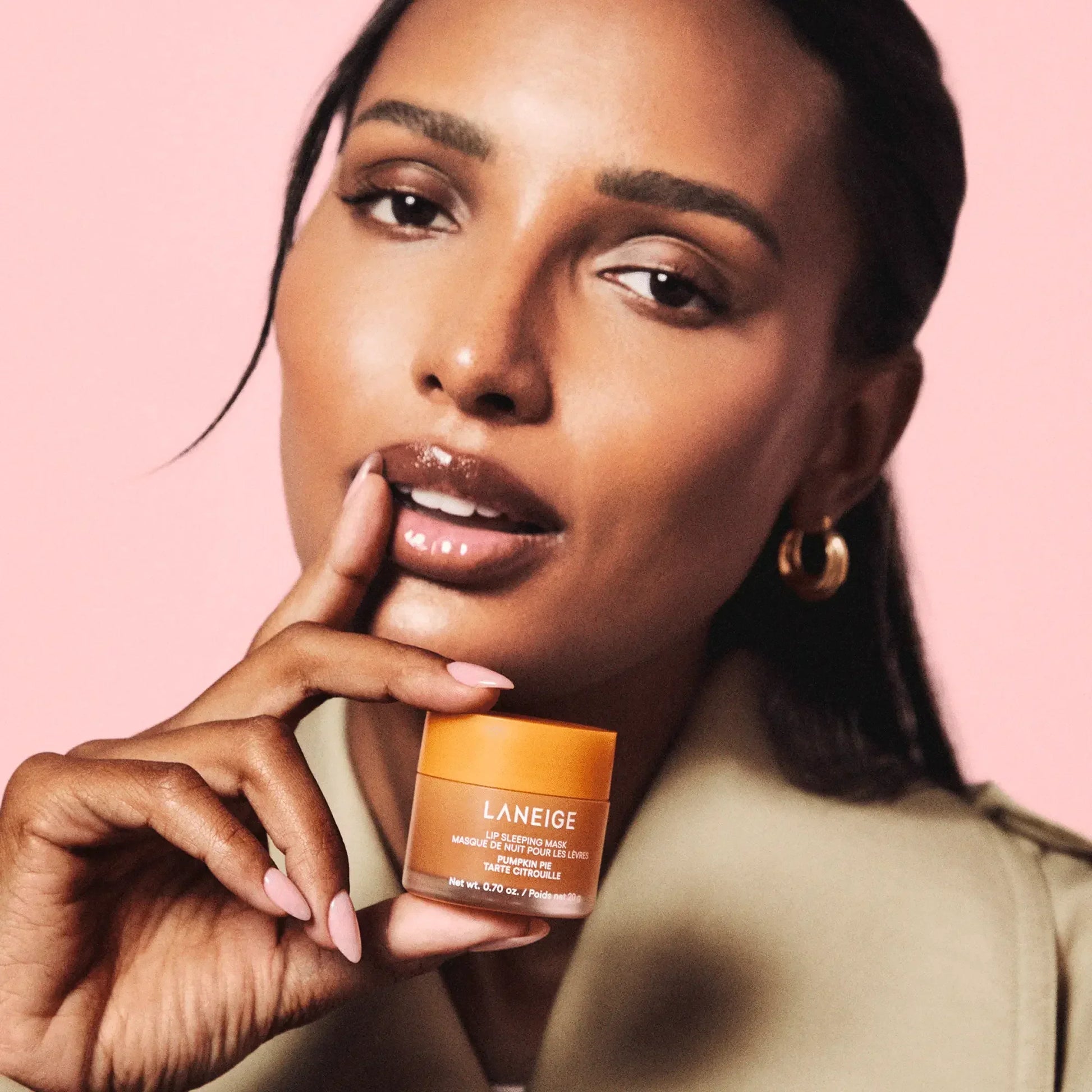 Woman with glossy lips holding Laneige Lip Sleeping Mask in orange jar against pink background.