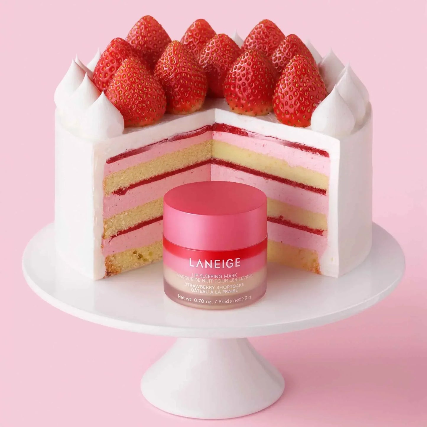 Laneige lip sleeping mask with a vibrant pink lid displayed in front of a strawberry shortcake with creamy layers on a white cake stand, set against a soft pink background.