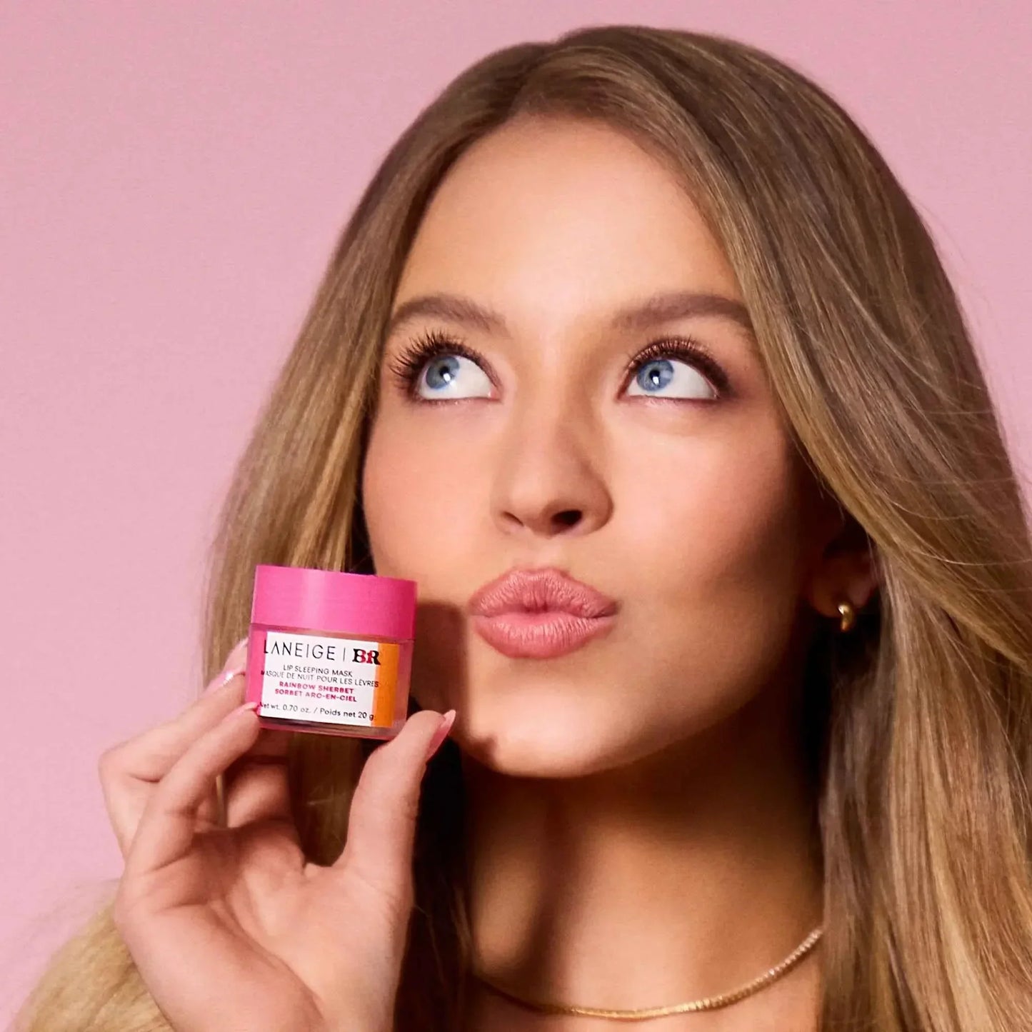 Woman holding pink Laneige lip sleeping mask, soft matte complexion, glossy lips, pink backdrop