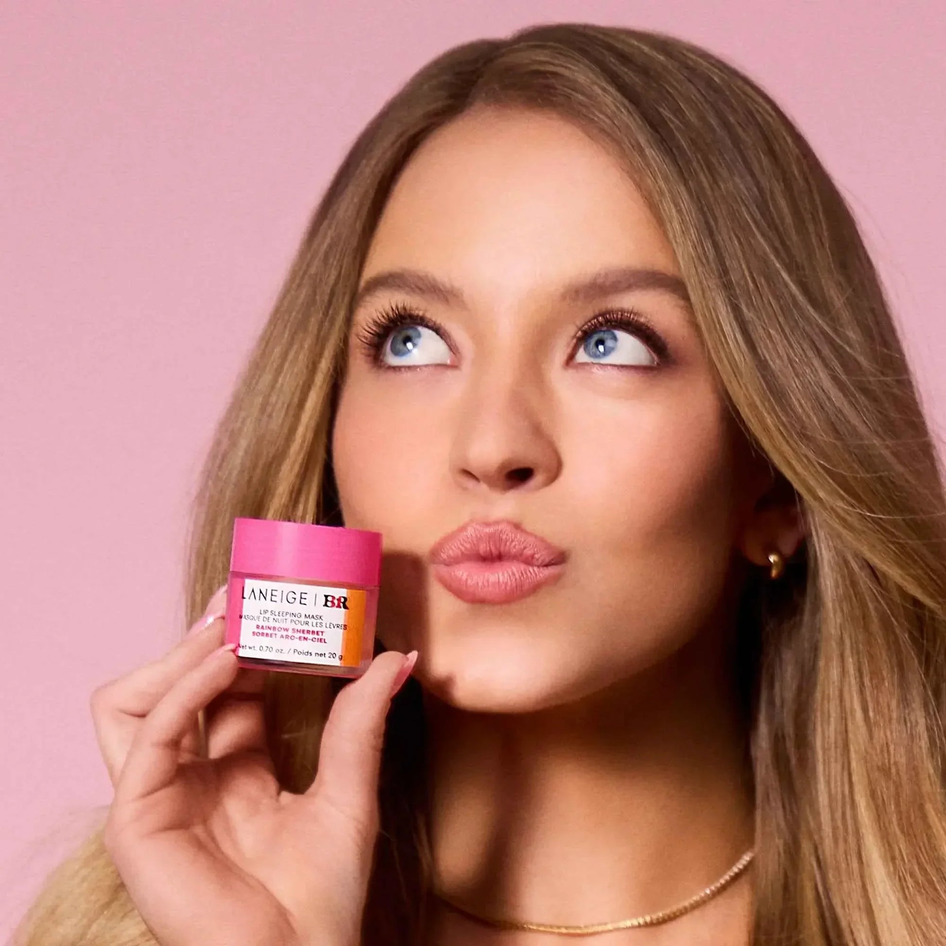 Woman holding pink Laneige lip sleeping mask, soft matte complexion, glossy lips, pink backdrop