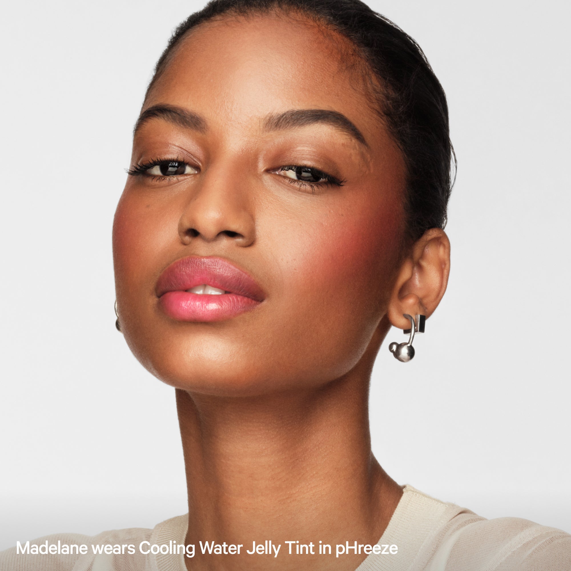Woman with dewy skin and pink jelly lip tint, wearing silver earrings, on white background.