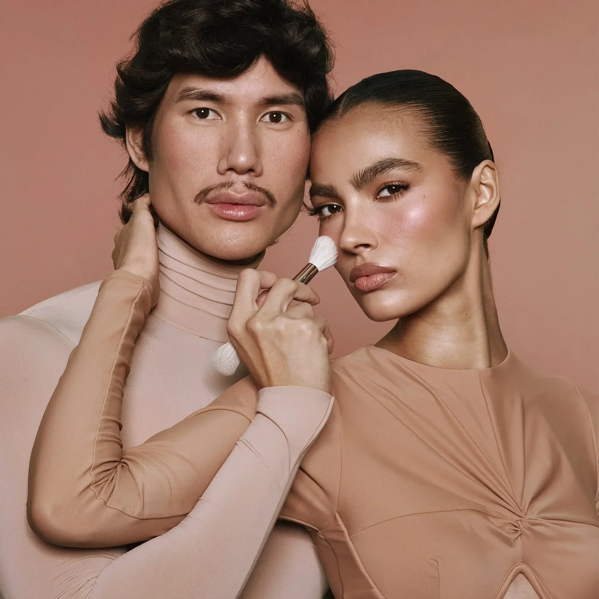 Two models in nude tones with flawless, dewy blush makeup and a soft makeup brush.