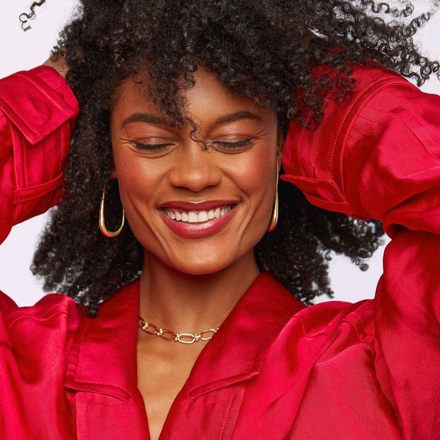 Woman with dewy, rosy blush and radiant skin wearing a red satin blouse and gold jewelry.