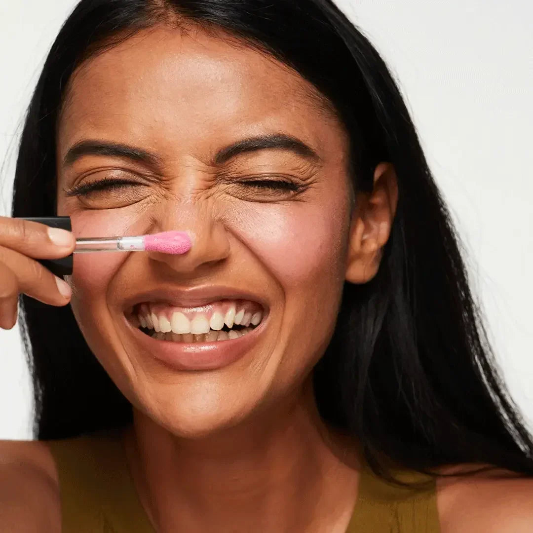 Smiling woman applying dewy pink blush with creamy texture to her cheek.