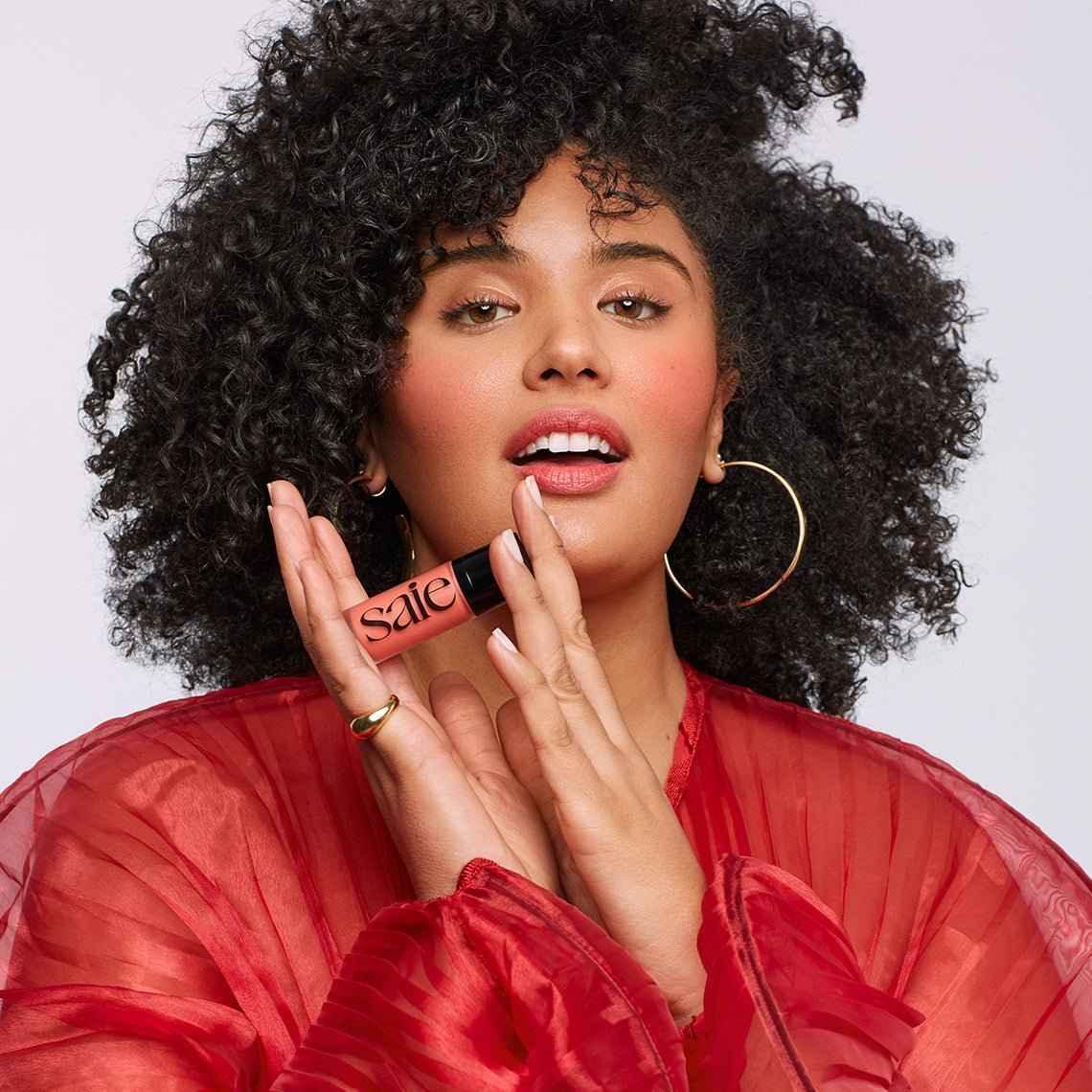 Woman with glowing skin wearing sheer red top, holding peachy dewy blush makeup.