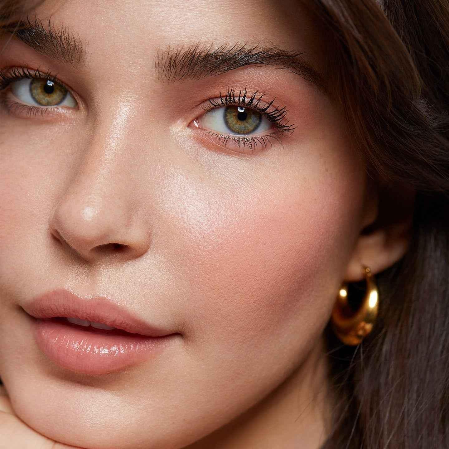 Close-up of woman with dewy skin, peach blush, soft pink lips, and gold hoop earring.