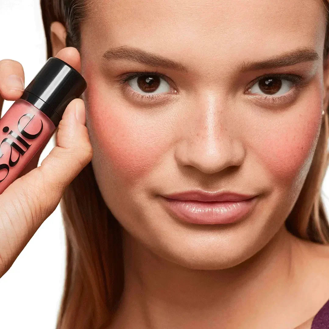 Model with radiant skin wearing pink dewy blush, holding blush bottle near cheek