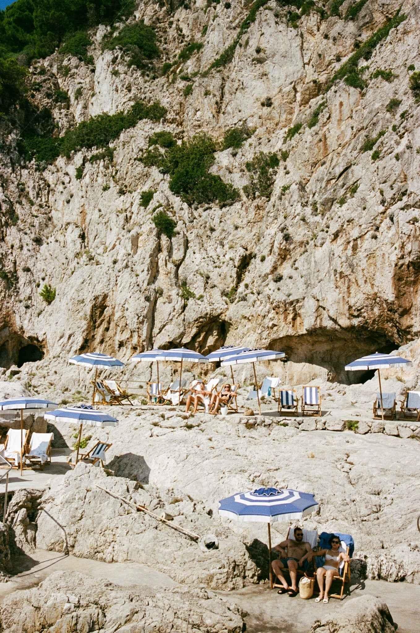Rocky cliffside beach with blue and white umbrellas, lounge chairs, and sunbathers relaxing in the sun.