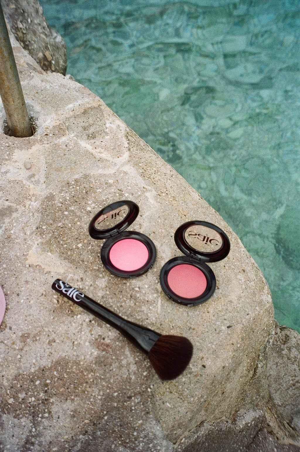 Two luxurious pink baked powder blush compacts with silky texture and a soft makeup brush on stone by clear blue water.