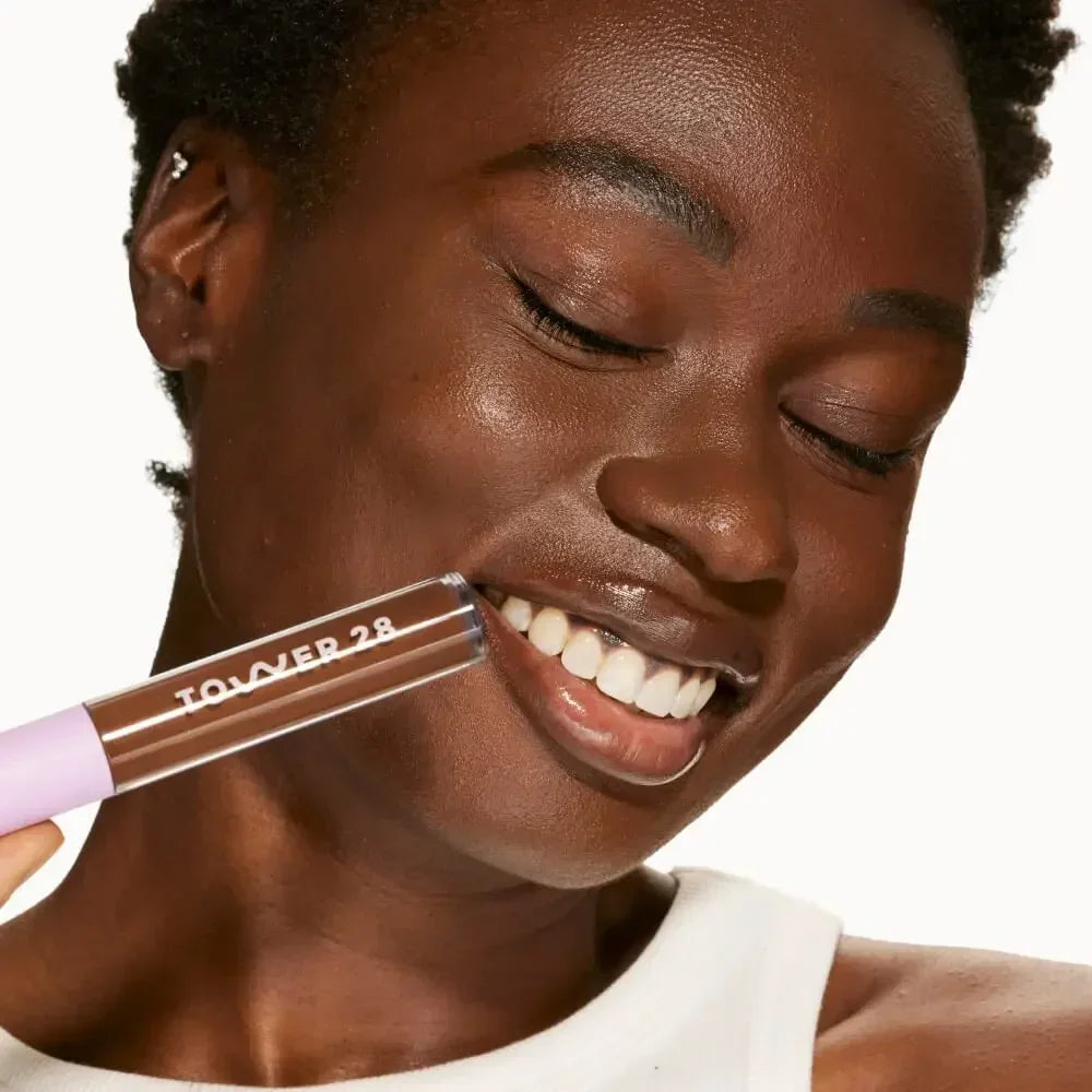 Smiling woman with radiant skin holding creamy brown serum concealer against her cheek
